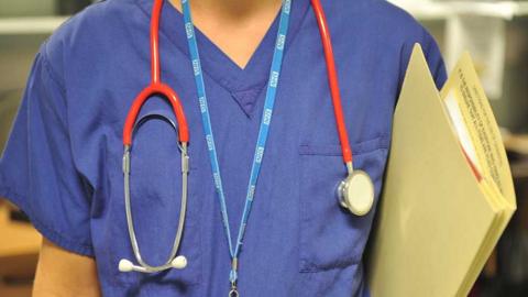 Generic image of the chest of a doctor in blue scrubs with a stethoscope and a blue NHS lanyard round their neck, holding a buff folder of medical notes under their left arm