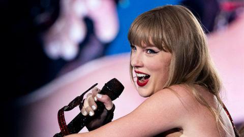 Taylor Swift, a female singer holding a black microphone in her right hand singing into it while smiling.