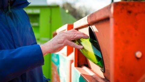 Person in blue jacket, hand visible putting a green glass bottle into a red recycling bin