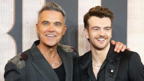 Robbie Williams has a huge smile and his arm around Jonno Davies who is also smiling. Both smartly dressed at a premiere event for Better Man.