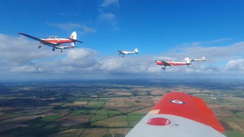 A view from the plane, and the planes fly in formation over Gloucestershire. The wing of the plane the photographer is in can be seen, and three light aircraft are in the sky. The sky is blue with white clouds and the fields are green and brown below. 