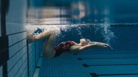 A woman in a white swimming hat and multicoloured costume, underwater in a pool. She is alone, on her back and her feet are touching the side - she is about to push off with her arms outstretched above her head