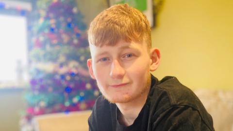 Teenage boy with ginger hair and black T-shirt smiling at camera, a decorated Christmas tree behind him in the corner.