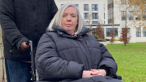 Karen James wearing a puffa jacket and sitting in her wheelchair on a grassy area in front of a block of flats