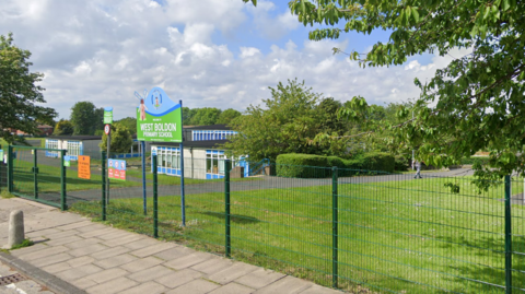 The entrance to the grounds of West Boldon Primary School, which sits behind a wire fence. Bright signage close to green, closed gates gives the name of the school.