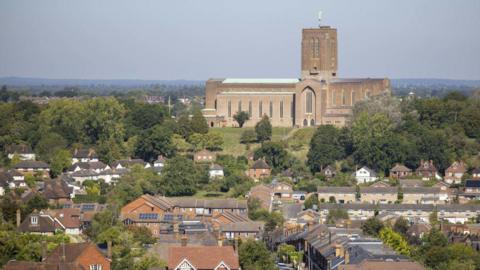 Guildford Cathedral is the largest building in a landscape that also includes houses and trees and a blue sky behind it. The cathedral is a modern-looking one with a square tower rising at its middle and long thin windows along its visible side