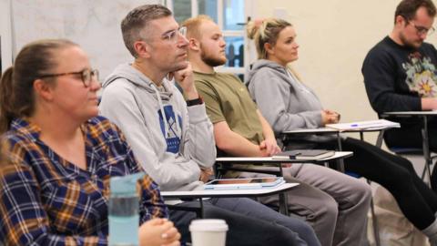 Five adults sat at desks in a classroom. They are all looking ahead. Three of the five are wearing glasses. 