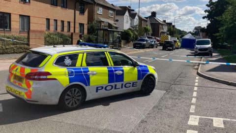 A police car is in the middle of a road. Part of the road has been cordoned off with white and blue police tape. A police officer can be seen and in the distance emergency service vehicles and a blue tent can be seen. 