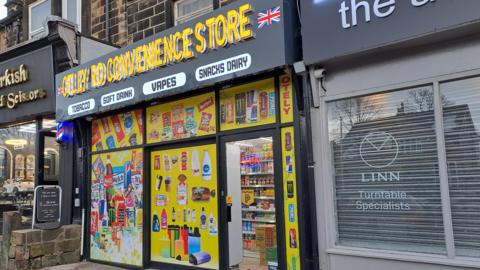 Otley Road Convenience Store. The shop has yellow posters in the windows with pictures of food and drink items.