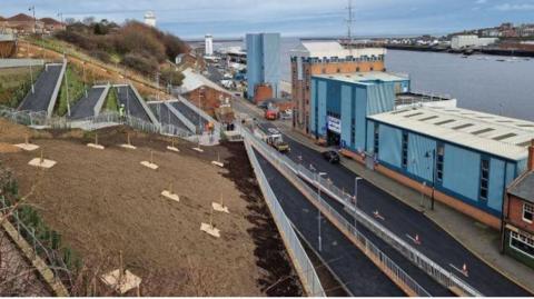 Aerial view of a zig-zag walkway running down a steep earth bank. A road runs along the bottom of the embankment. In front of that is a long blue building which runs behind the River Tyne.