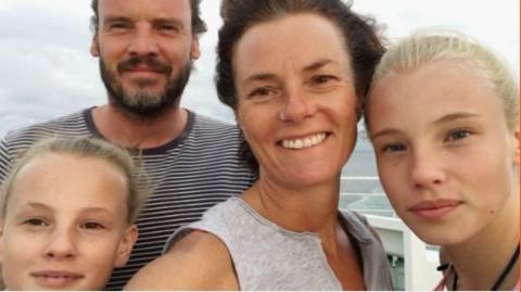 Kate takes a selfie with her two blonde daughters and partner Ian, who has a brown beard and is wearing a striped t-shirt. Kate has short brown hair and a nose ring. She smiles broadly at the camera, wearing a grey top.