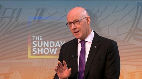 John Swinney wears a black suit jacket with saltire pin, white shirt and purple tie. He stands in front of a screen with the Sunday Show logo in orange, blue and yellow. He wears black thin-rimmed glasses and gestures with his right hand as he speaks. 