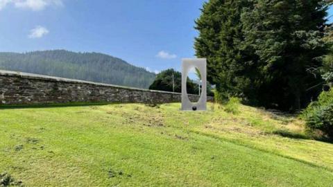 A steel modern art sculpture, which is a rectangle with an oval shaped hole, standing on some grass. It is placed next to a low stone wall, with a tree-covered hill in the background.  