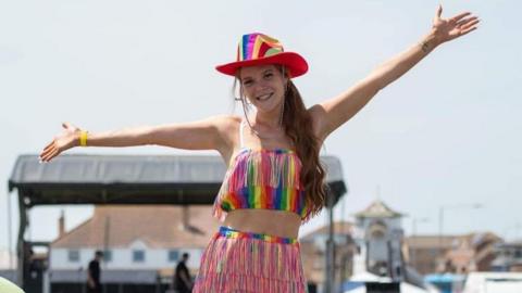 A woman wearing a rainbow top, skirt, and hat with her hands in the air