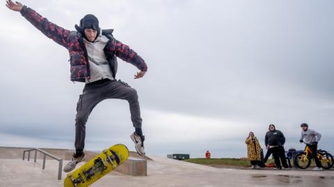 A young man doing a trick with a skateboard at the new Maryport skatepark. There are three bystanders watching, including a man on a BMX. 