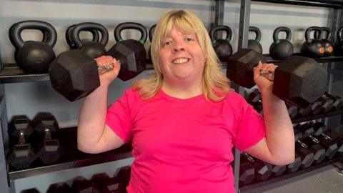 Aisling McCooey holding two weights in the gym. She has blonde hair and a pink tshirt. She is smiling.