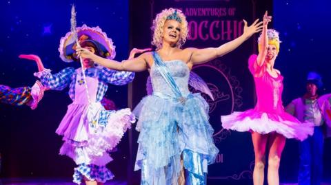 A blond drag queen wearing a blue sparkly dress and a tiara stands with her arms outstretched on a stage. Dancers perform on either side of her. 