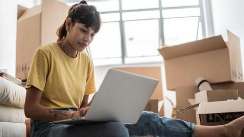 A young woman looks at a laptop surrounded by cardboard boxes