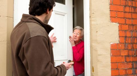 An elderly woman answers a door to a man. 