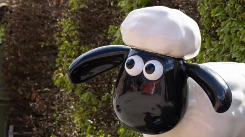 Model of animated character Shaun the Sheep. A close-up of his head shows him wearing a white hat. He has a black face and large eyes and his body is white