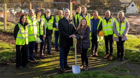 Representatives from Aspire Housing, Homes England, Newcastle-under-Lyme Borough Council and Keon Homes. There are several people wearing fluorescent yellow jackets, while a man and a woman at the front are holding a shovel. There is construction fencing behind the group.