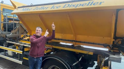 Mike Zeller stands in front of the yellow gritter which bears the name Mike Zeller Ice Dispeller on its side. Mike is pointing at the name and smiling.