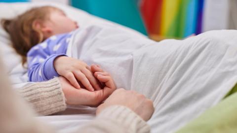 A stock image showing a young child in a bed. The girl is holding an adult's hand.