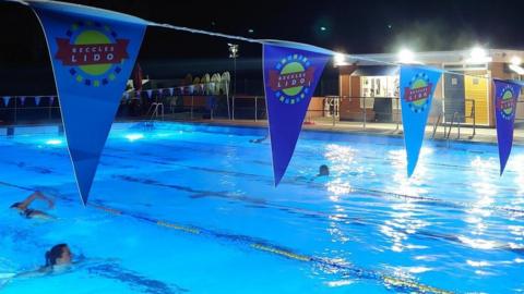 Beccles Lido at night. There is blue bunting across the pool and several people are swimming.