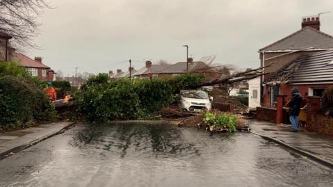 A fallen tree lies across a road. To the left is a man in a high visibilty jacket and to the right a white car, partially covered by the fallen tree, the top branches of which appear to have damaged the roof of a single-storey building