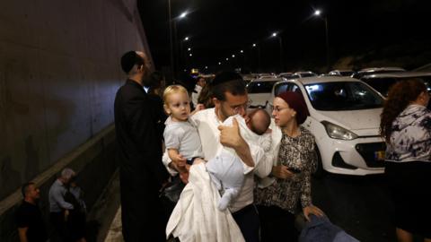 A man holds children as people take cover as air raid sirens sound across Israel during an Iranian ballistic missile attack, in central Israel (1 October 2024)