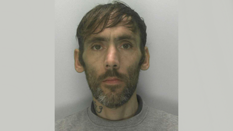 A police mugshot of Matthew Cross. He is 45 years old, with a sweeping brown fringe and full brown beard which is turning grey on the chin area. He is wearing a grey sweatshirt and has a symbol tattoo on the right side of his neck. He is standing against a grey background.