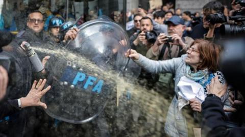 A woman being sprayed with pepper spray by a police officer behind a riot shield