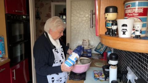 Val standing in her kitchen wearing a Fred apron. She is pouring flour through a sieve into a pink mixing bowl. On the shelves and counters surrounding her there are many Fred memorabilia items.