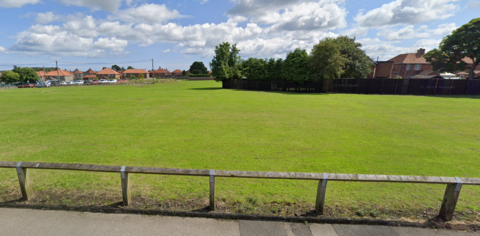 A low wooden fence is in the foreground with a large playing field behind fringed with a number of houses 