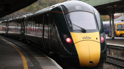 GWR train at a train station. The train is yellow at the front with red headlights and a dark green body. The platform is empty.