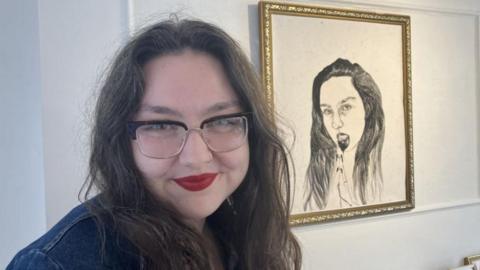 A woman with long dark hair, nose ring and rectangle glasses, wearing red lipstick. She is standing in front of a gold-framed sketch of a woman, which is in the background