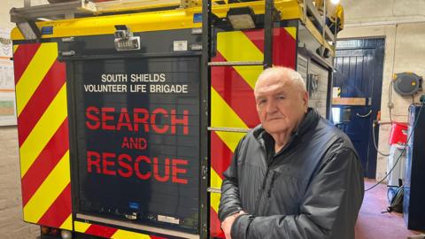 Tom Fennelly standing in front of the South Shields Volunteer Life Brigade rescue vehicle, which is red, yellow and blue. 