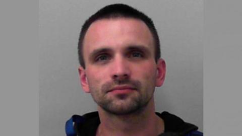 A police mugshot of Dominic Scribbins. He is wearing a black hooded jacket and standing against a grey background. He has short dark hair, a short beard and blue-grey eyes. He is looking directly into the camera.