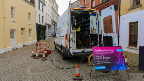 The back of an van for fibre broadband that has its doors open and wires coming out of it. There is a sign that reads full fibre is coming in front of the open doors and traffic cones around the van.