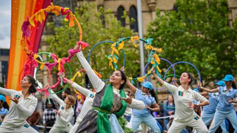 A group of women, some in cricket outfits, hold hula hoops above their heads while dancing.