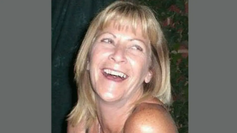 Jean Hanlon looking slightly off camera. She has shoulder-length blonde hair and is laughing. Her top row of teeth are visible. She has light-coloured eyes. Only her shoulders, neck and head can be seen.