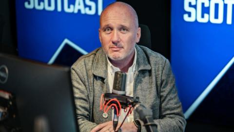 Martin Geissler sits at a microphone in a radio studio, with scotcast logo in background, wearing grey shirt with grey stubble on face.