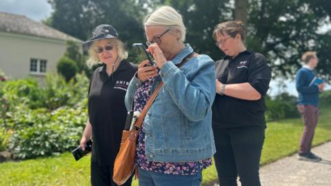 A woman in a blue denim jacket and carrying a light brown handbag looks closely at her phone while walking around the gardens at Saltram. Two women from the Royal National Institute of Blind People with black polo shirts with RNIB branding on it are behind her.