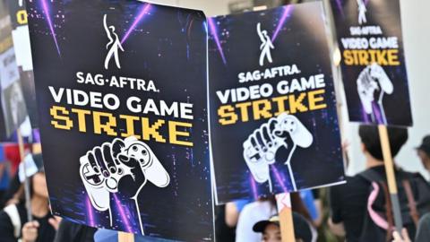 Three placards being held in the air with a picture of a fist clenched around a gaming controller and the text "Sag-Aftra on strike!". 