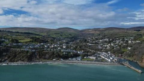 An aerial view of Laxey Bay featuring a turquoise sea and green hills in the background. White buildings scatter the landscape.