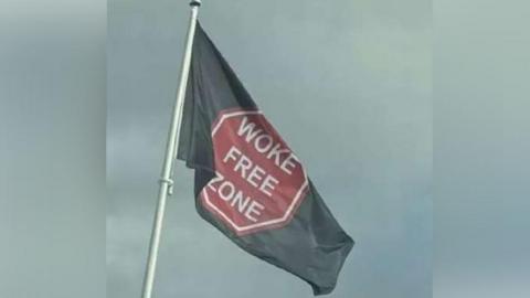 A black flag blows in the wind. It bears the slogan 'woke-free zone'. The words sit in a red and white octagon