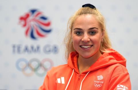 Olympic rower Becky Wilde smiles at the camera with a large Team GB logo in the background. She is wearing a branded Team GB red hoodie which also has an Adidas logo on it