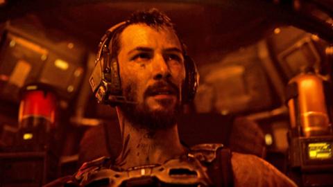 A battle-scarred man sits inside the futuristic cockpit of a robot craft, illuminated in a dim orange light emanating from the controls. He wears a communications headset and a metal harness.