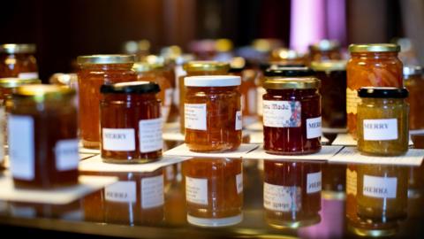 View of a number of jars of marmalade, ranging in colour from dark to light orange. All are labelled and are resting on a mirrored surface to reflections can be seen.