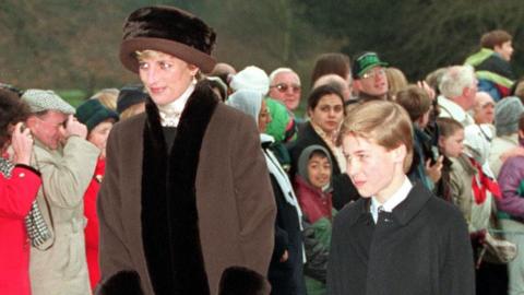 Princess Diana wearing a brown hat and coat with dark brown fur trim and a young Prince William wearing a shirt and tie and black overcoat walk past crowds lining a path towards church on Christmas Day.